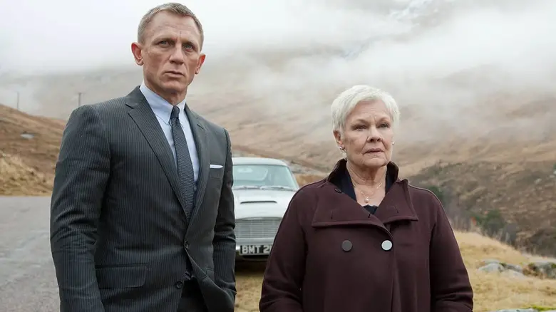 Daniel Craig as James Bond standing next to Judi Dench's M in front of the Astin Martin DB5 in Skyfall