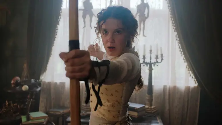 Enola Holmes (Millie Bobby Brown) looks surprised while holding a bow in Enola Holmes