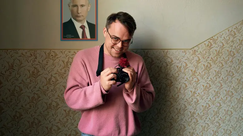 Pavel Talankin flashes a mischievous grin and holds a camera in front of a picture of Vladimir Putin in Mr. Nobody Against Putin