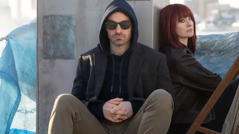 Charlie Cox as Matthew Murdock wearing a hoodie and sunglasses and seated next to Deborah Ann Woll as Karen Page undercover in a red wig in Daredevil: Born Again