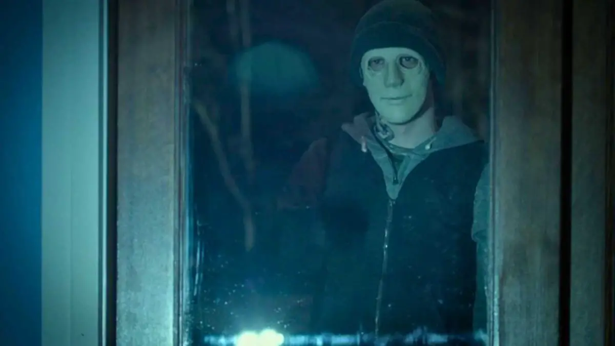 A man in a zipped up hoodie with a white ceramic mask and a beanie stares inside a darkly lit house.