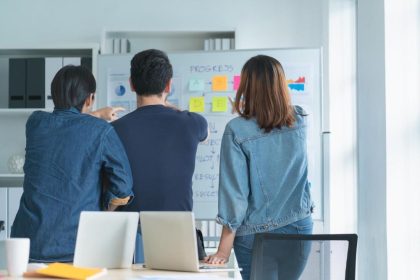 Três colegas analisando uma estratégia de negócios e um plano de progresso em um quadro branco durante uma reunião de equipe.